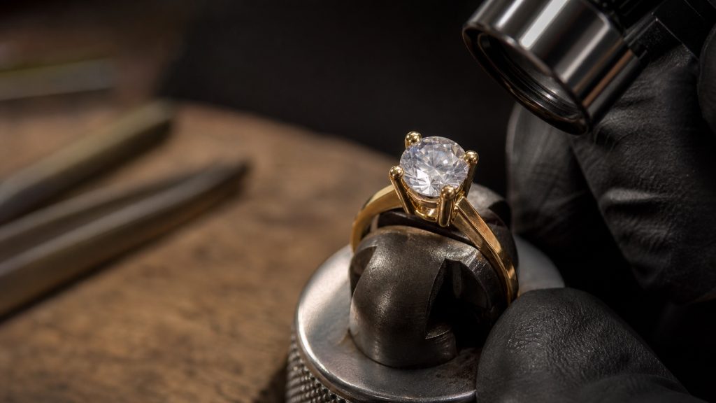Anel de ouro com pedra redonda em garras sendo inspecionado sob microscópio na bancada de cravação